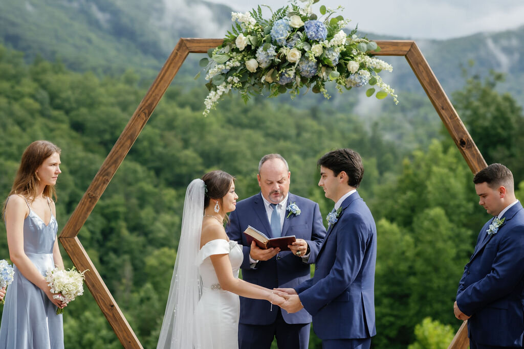 wedding photographers gatlinburg tn captures outdoor ceremony with bride and groom holding hands and listening to their officiant with the Smoky Mountains in the distance captured 