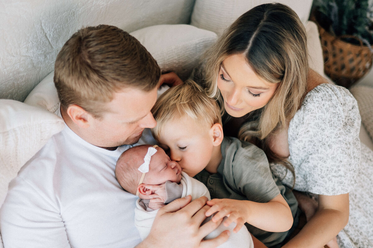 family of four with newborn on dad's chest with brother kissing her and mom looking on