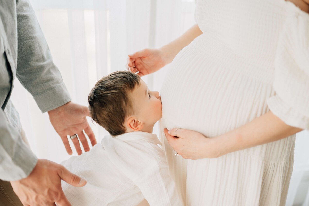 toddler kissing his pregnant mother's belly with dad's hands supporting beside him