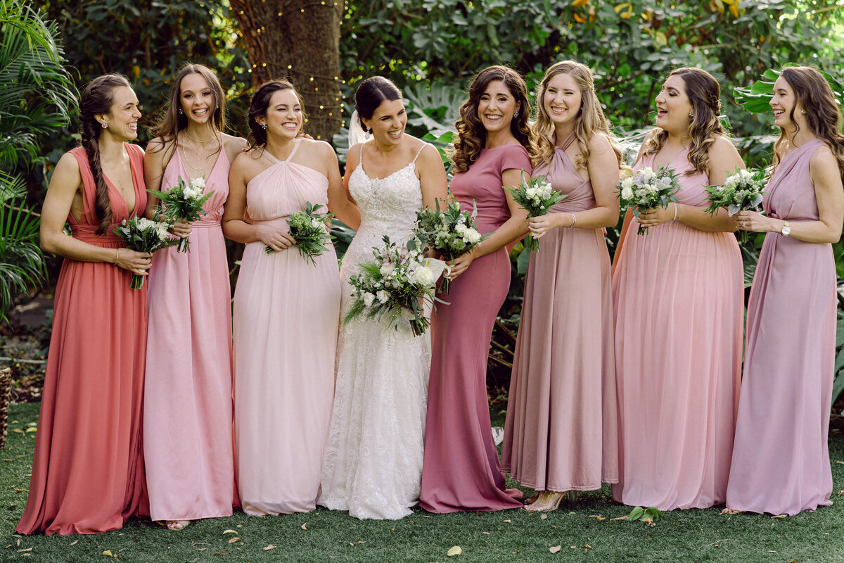 A joyful bride with her bridesmaids in pink dresses, sharing laughter in a lush garden, captured by Rodrigo Varela Photography.
