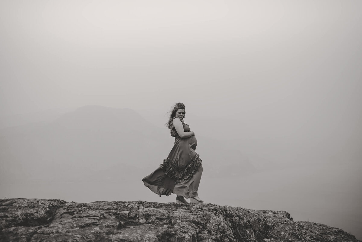 A pregnant woman stands on the edge of a cliff in this black and white portrait taken in Kamloops by Brittany Danielle. The wind is blowing her hair and  dress while she holds her belly.