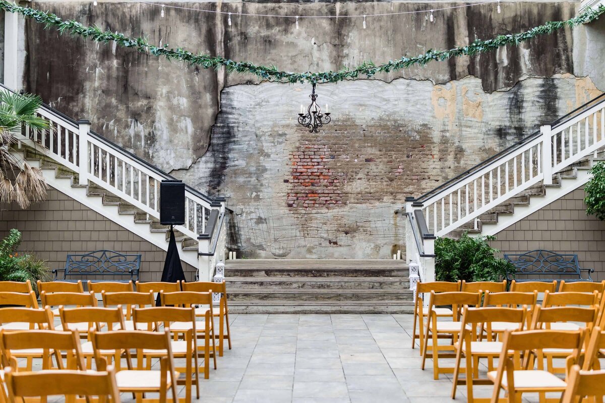 Rows of wooden chairs face a small stage with steps, flanked by two white staircases. A weathered brick wall, string lights, greenery garland, and a black chandelier decorate the outdoor venue.
