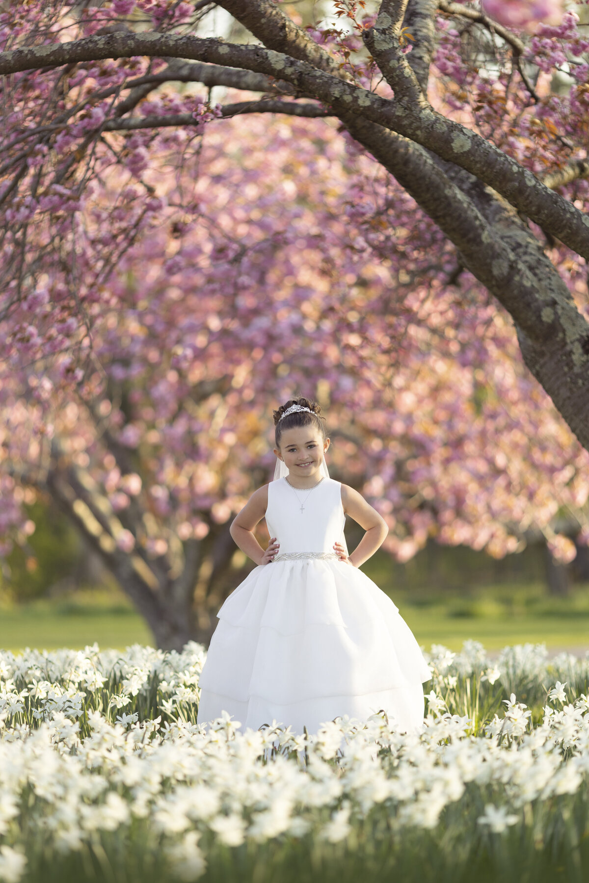 A proud young girl wearing a long white silk dress with hands on her hips stands in a field of flowers