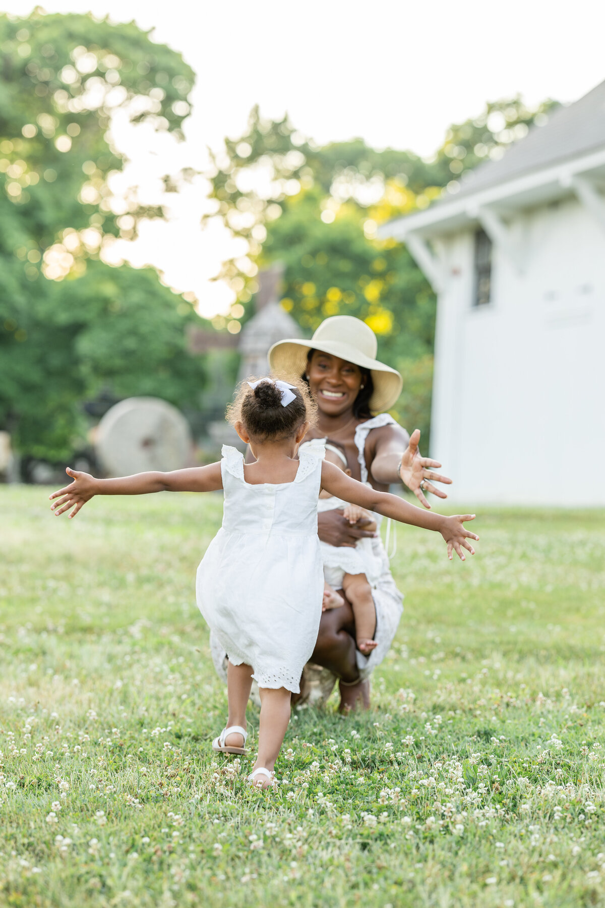 A young girl in a white dress runs with open arms towards her smiling mother, who is crouched  down with her arms wide open, ready to embrace her. 