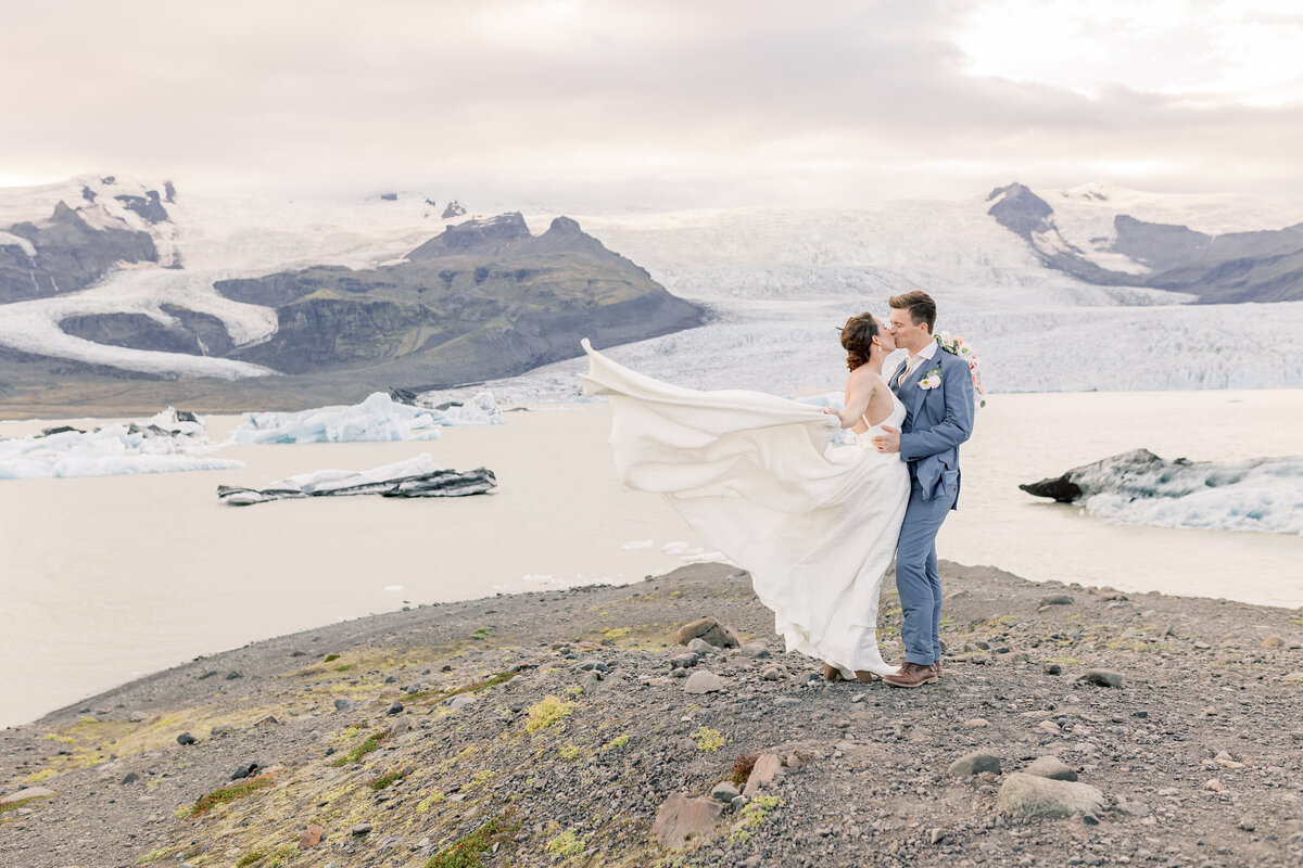 Bride's dress blowing in the wind as she kisses her groom in front of a lake of glaciers near glacier lagoon in Iceland.
