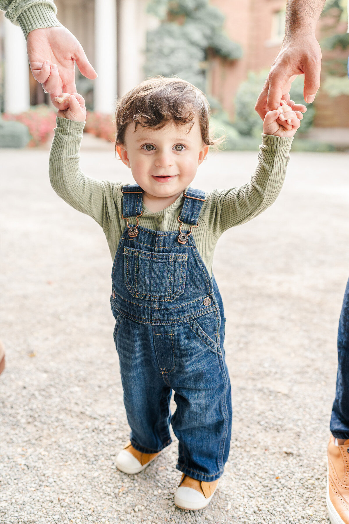 toddler boy in overalls holding his parents' hands