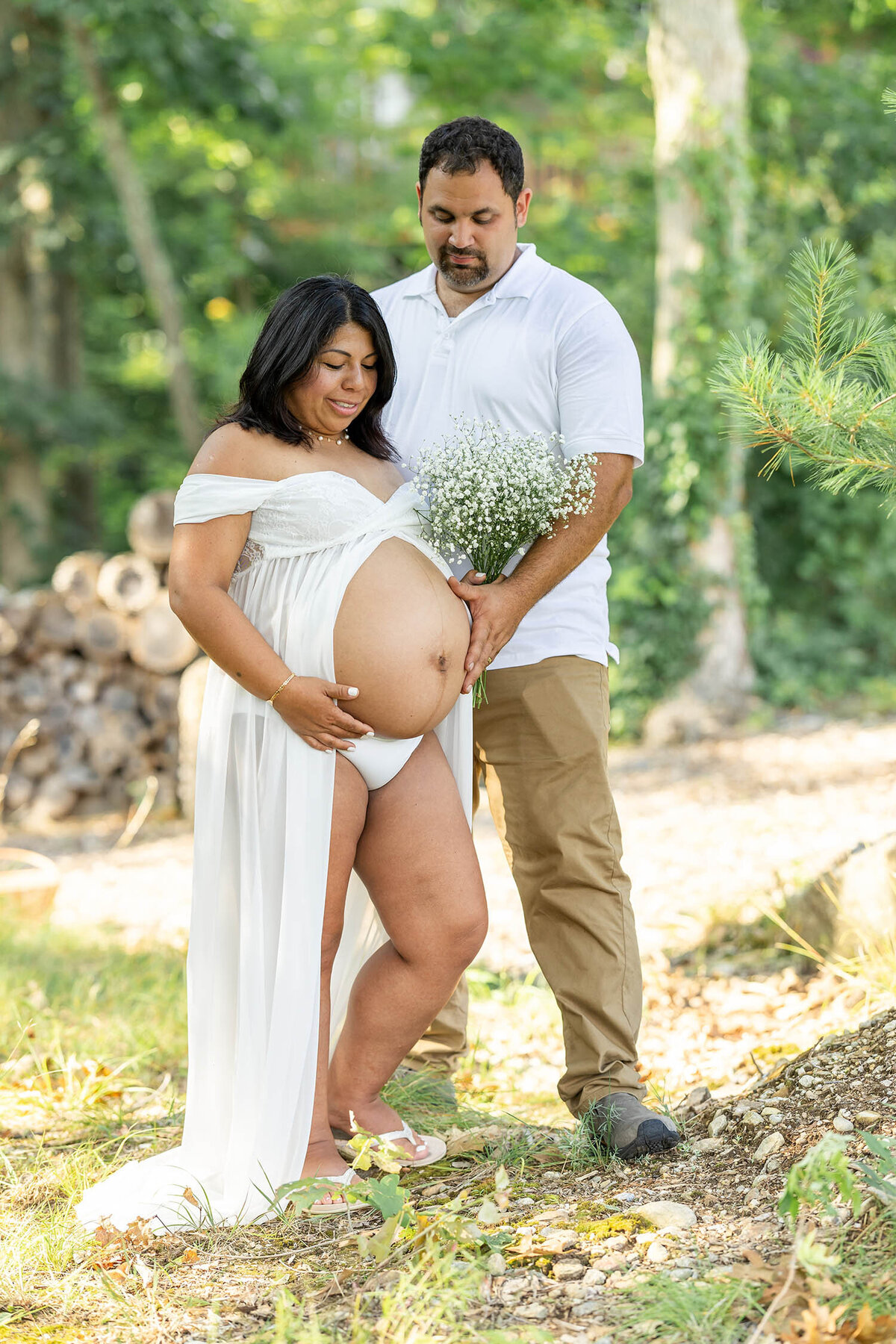 Pregnant woman wearing an open belly white dress and a man standing next to her as they both look down at her bump and smiling during their maternity session in Connecticut 