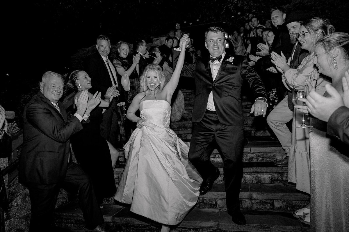  Bride and groom celebrate during their grand exit, running down the stone steps while guests clap and cheer on both sides at Old Edwards Inn in Highlands, North Carolina.