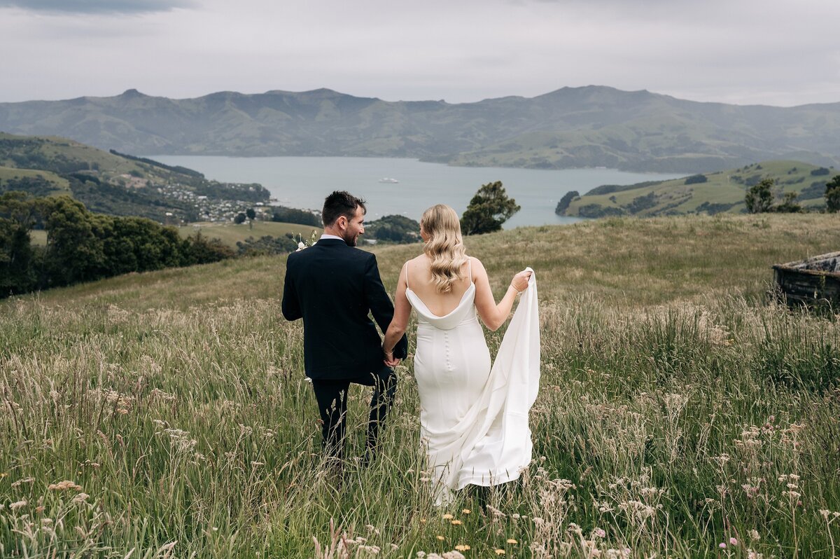 bride and groom walking away from camera in field looking at the view over akaroa harbour in new zealand on a cloudy day
