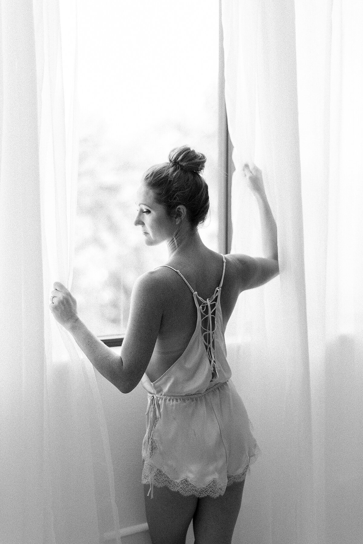 Black and white in studio boudoir photo of a woman dressed in a lace romper while she holds sheer drapes while standing by the window.