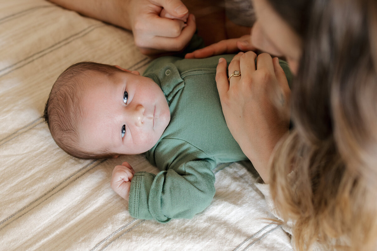 Longmont newborn looking at mom on bed while mom holds babys belly and dad holds babys hand