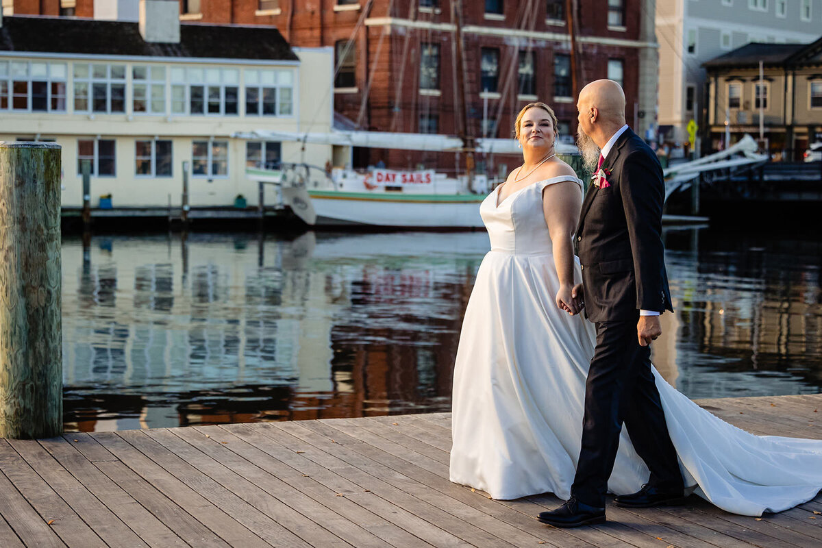 The bride and groom stroll along a waterfront boardwalk, with boats and reflections on the water behind them