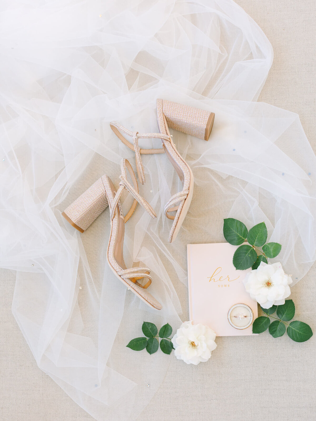 Nude high heels on white tulle fabric, next to a pink "her vows" book, wedding ring, and white flowers with green leaves.