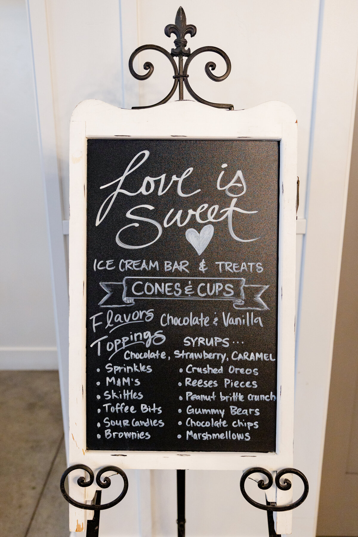 A charming blackboard sign with hand-lettered calligraphy reading "Love is Sweet," showcasing an ice cream bar with flavors, toppings, and syrups available for wedding guests. A whimsical touch to a beautifully curated reception. 