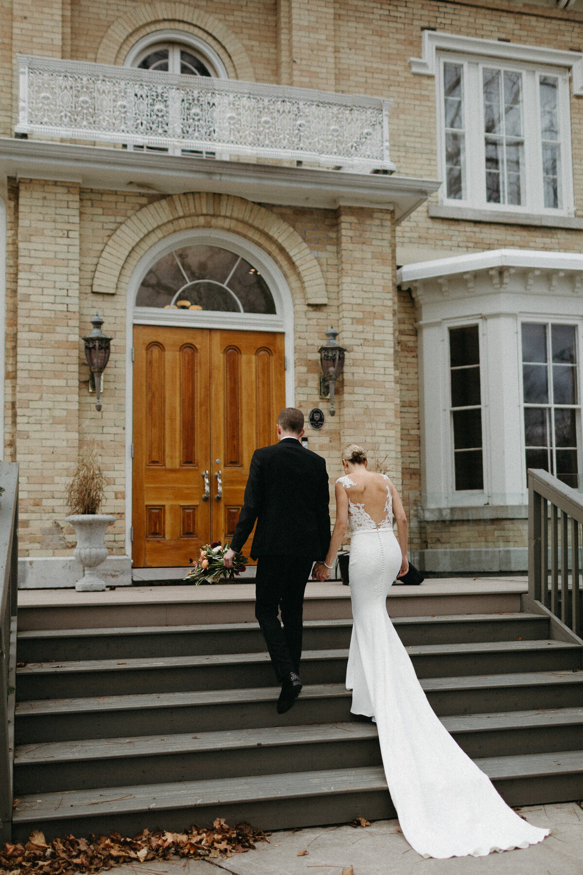 A couple in wedding attire ascends the stairs towards a grand wooden door of a brick building at Cape Picton. The bride's white dress and the groom's black suit evoke timeless elegance, perfectly captured by their Muskoka wedding photographer.