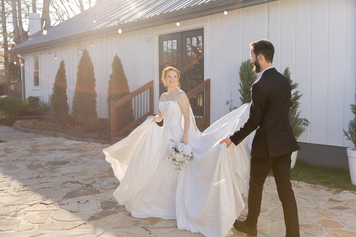 A groom holds the train of his bride's dress as the walk away from the camera. Behind them is  white house.