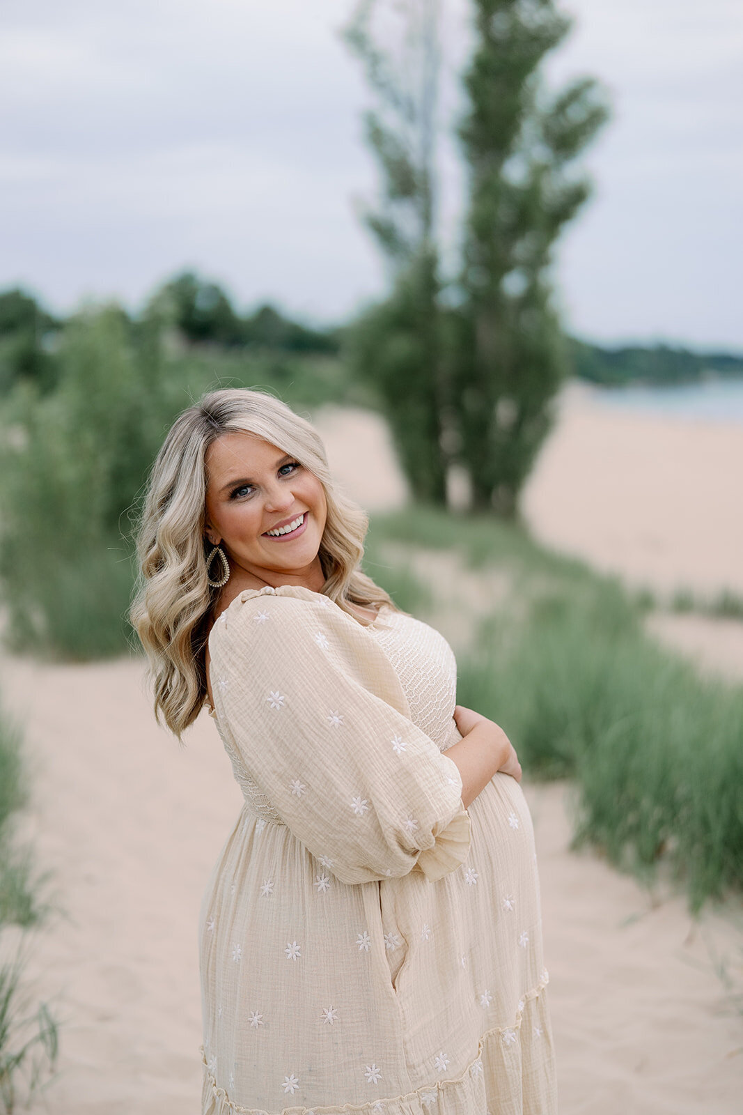 Pregnant mother smiling and holding her belly in the grassy dunes at South Haven North Beach during a maternity photography session.