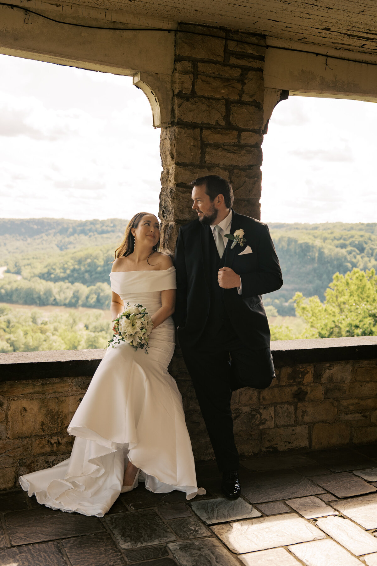 A bride is sitting on a ledge with the Ohio River in the background and a groom is leaned up against a stone pillar next to her. 