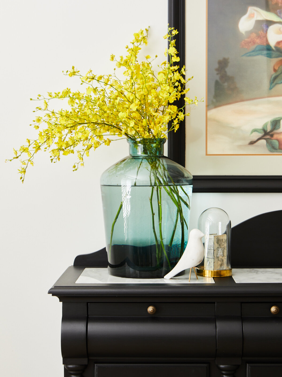 A close-up of a styled black console table in an Eynesbury home, featuring a large teal vase with vibrant yellow flowers, a small white bird figurine, and a decorative glass cloche.