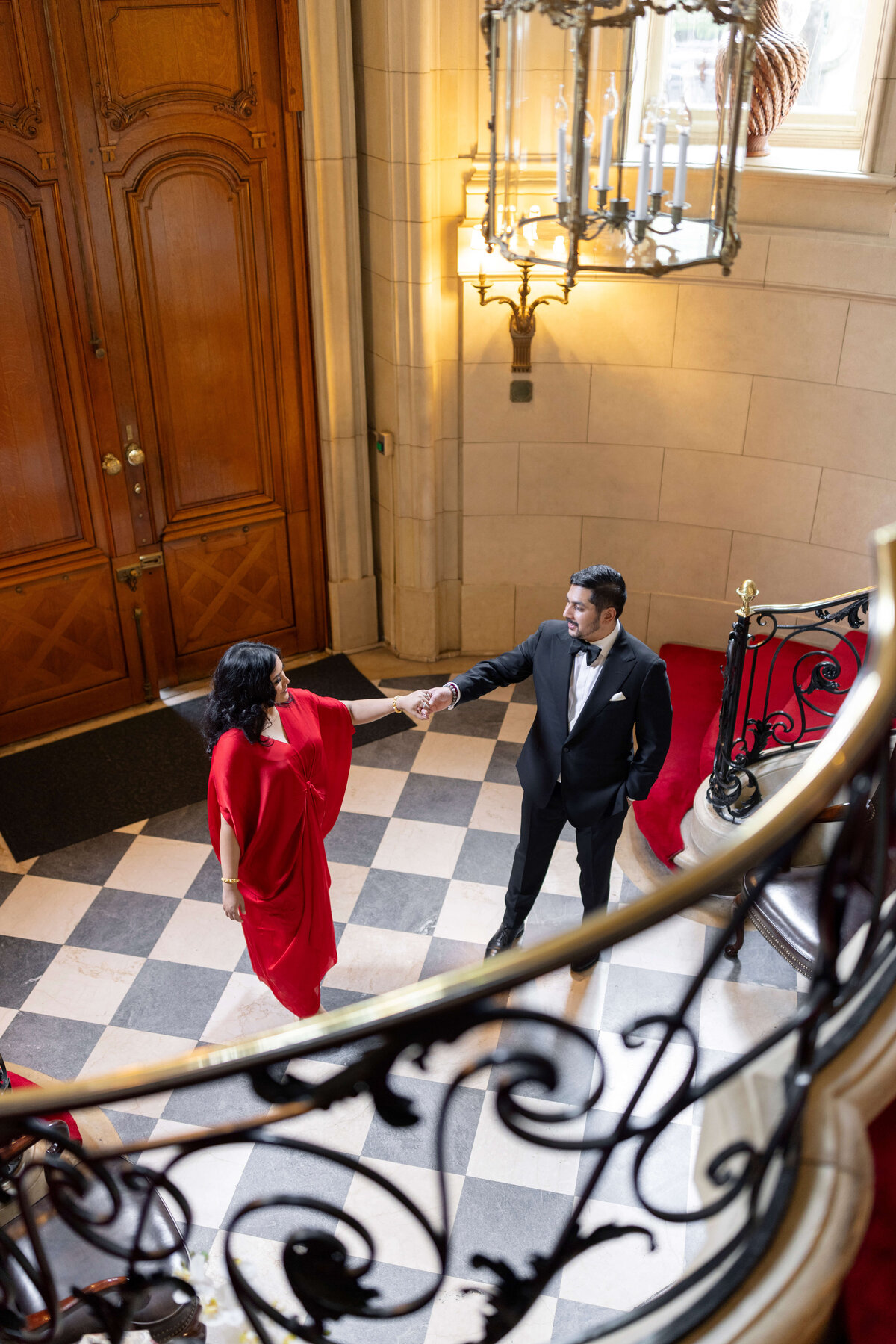 A couple elegantly dressed, with the woman in a red gown and the man in a tuxedo, stand on a checkered floor by a grand staircase with intricate railings. Theyre about to descend, with a large chandelier overhead and wooden doors in the background.