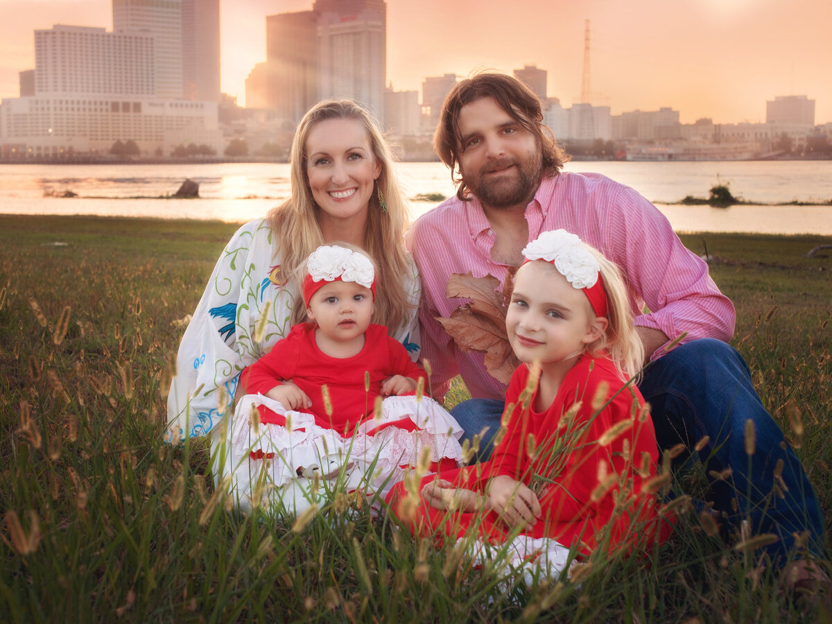 Family of 4 sitting on the levee in Algiers Point with the New Orleans skyline in the background.   Girls are wearing red dresses and headbands.  Mom is wearing a boho dress.  Dad is wearing a red gingham shirt.