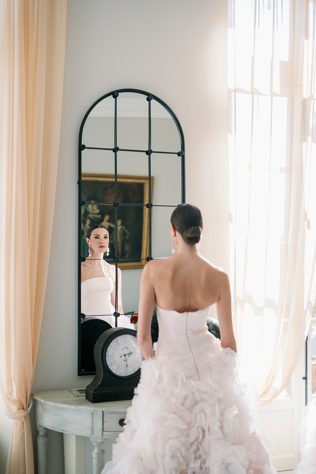 Bride looking into tall arched mirror with soft window light in a French bridal suite at Château de Tourreau.