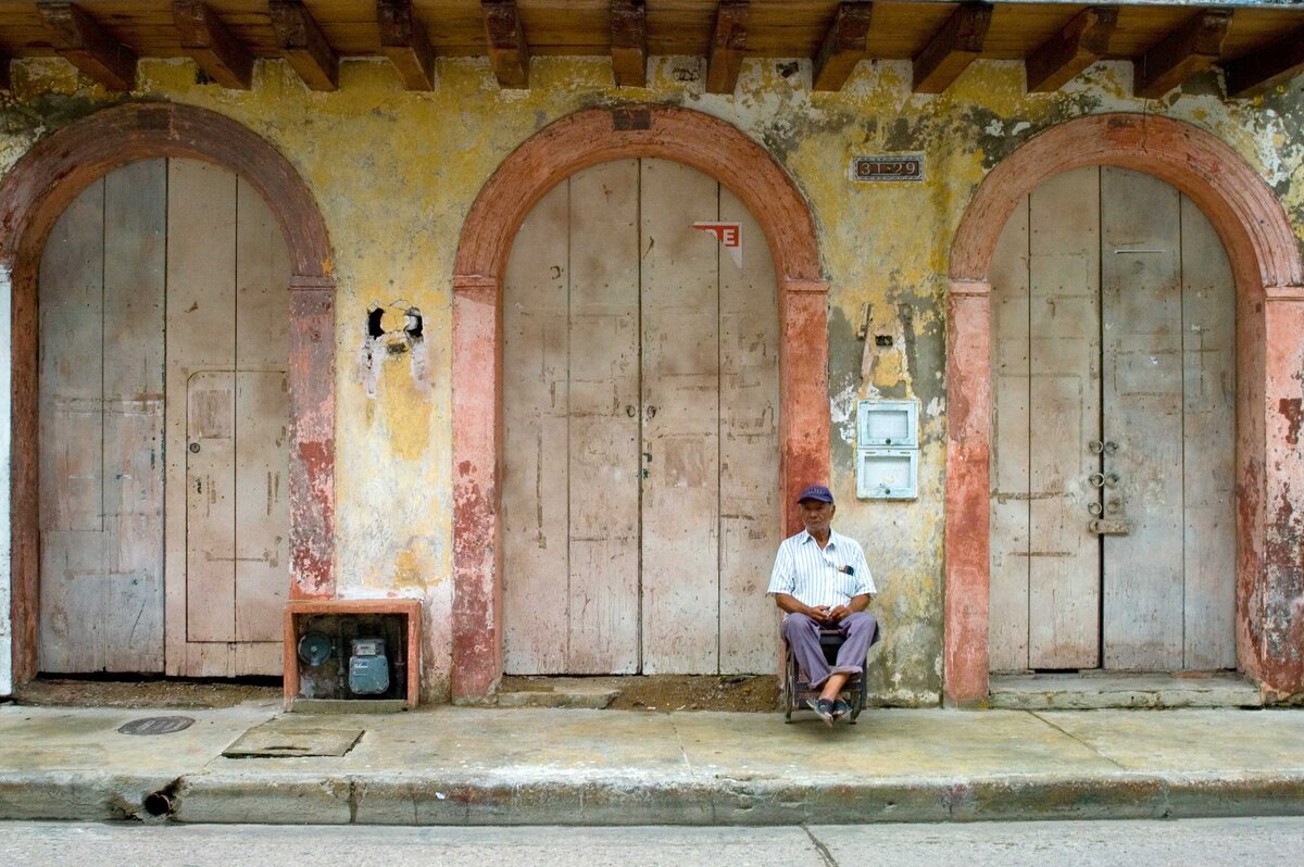A man leans back on a chair in front of a series of doorways in a street scene in Cartagena. This artistic journalistic image depicts the streets in the old part of town.