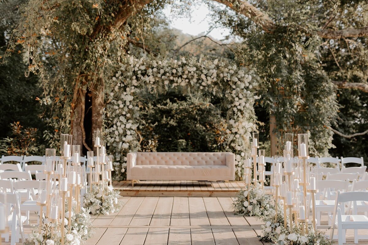 Luxury wedding ceremony setup with white and greenery filled arch, candles lining the aisle inside flower arrangements at the Bowery House and Gardens in Houston TX.