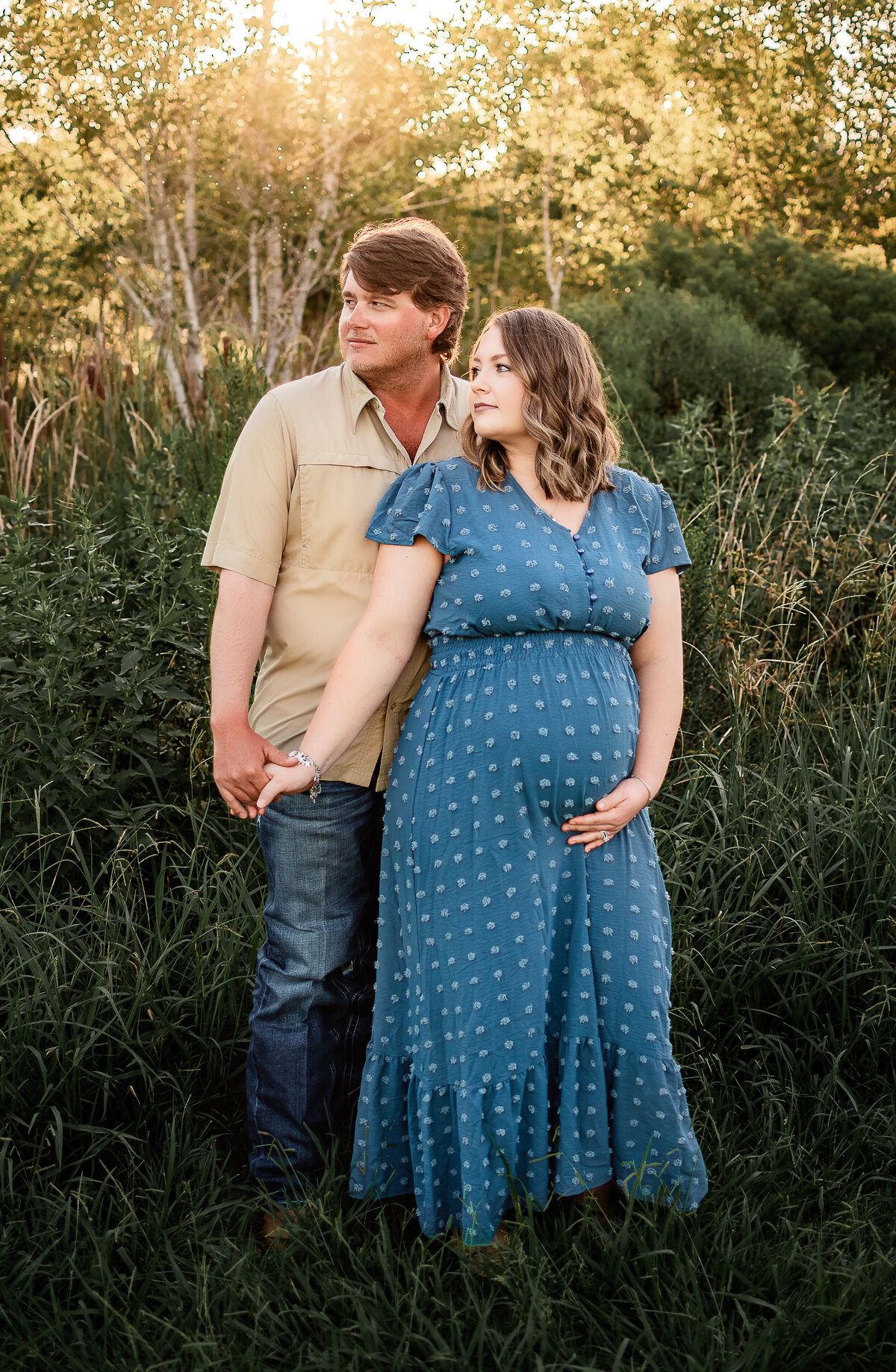 A pregnant couple holds hands and looks into the distance while the sun shines behind them.
