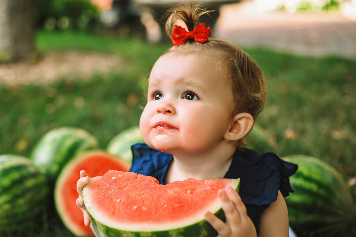 Little girl looks away as she holds a slice of watermelon below her chin