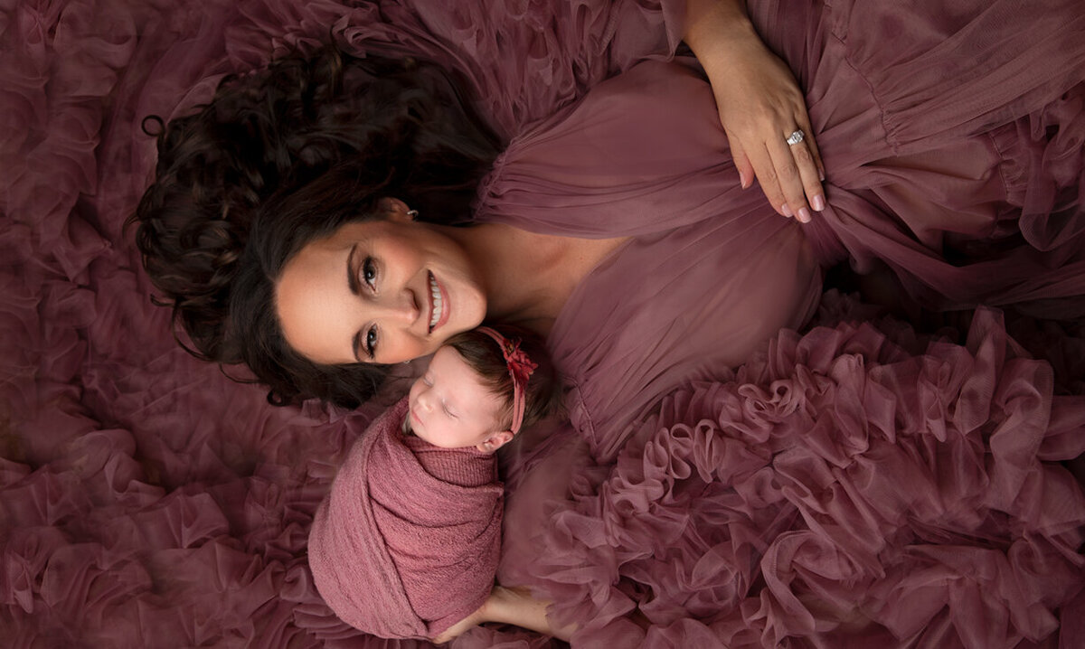 A loving mother cradles her newborn baby while posed gracefully on the floor, dressed in a stunning mauve fluffy couture gown that envelops both mom and baby. This elegant portrait, captured at Little Legacies Studio in Phoenix, Arizona, highlights the beauty of motherhood and the tender connection between mom and baby.
