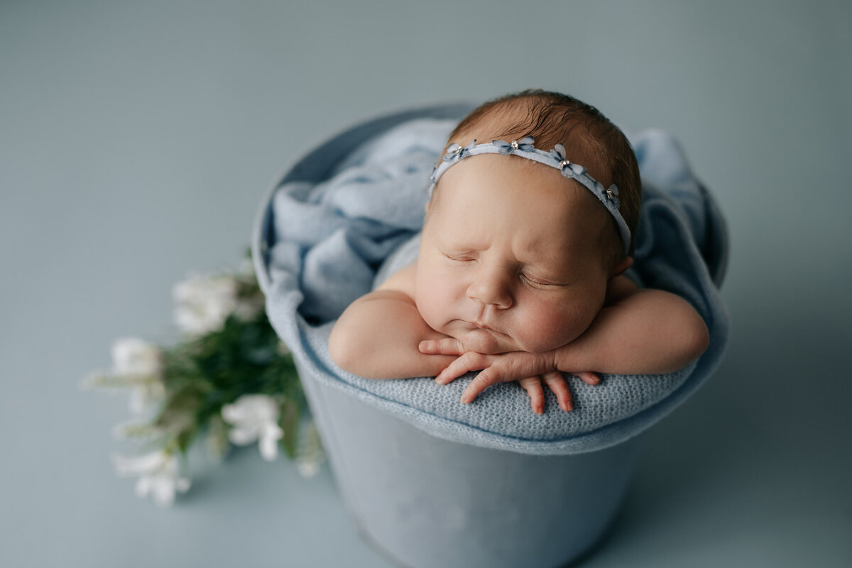 Studio newborn photography - baby girl sleeping in metal bucket. Baby is on a blue blanket with a petite blue floral headband. Flowers placed beside bucket.