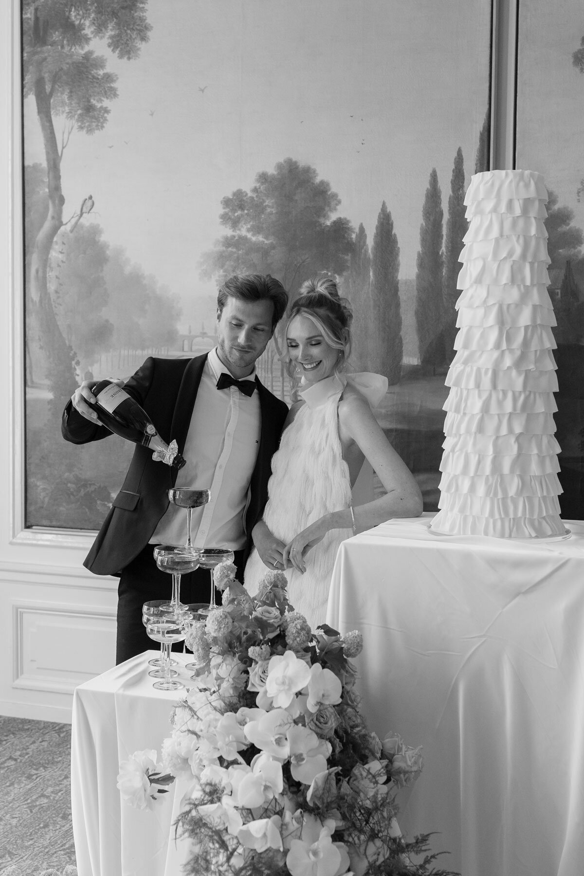 Luxury 3-tier white wedding cake with ruffles with couple drinking champagne at a high-end wedding.