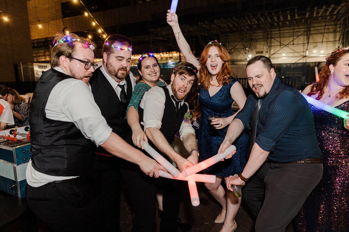 A groom and his wedding party all touching their light up foam sticks together in celebration during his wedding reception at the Pegasus City Brewery in Dallas, Texas. The groom is on the far left and is wearing his wedding suit (minus the jacket) and a light up crown. His wedding party has on similar light up crowns and are all cheering in celebration.