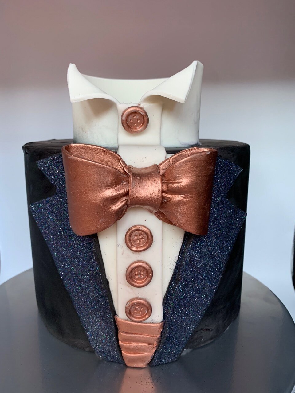 Single tier tuxedo cake design with shiny black labels, bronze colored bow tie, cummberbund, and buttons