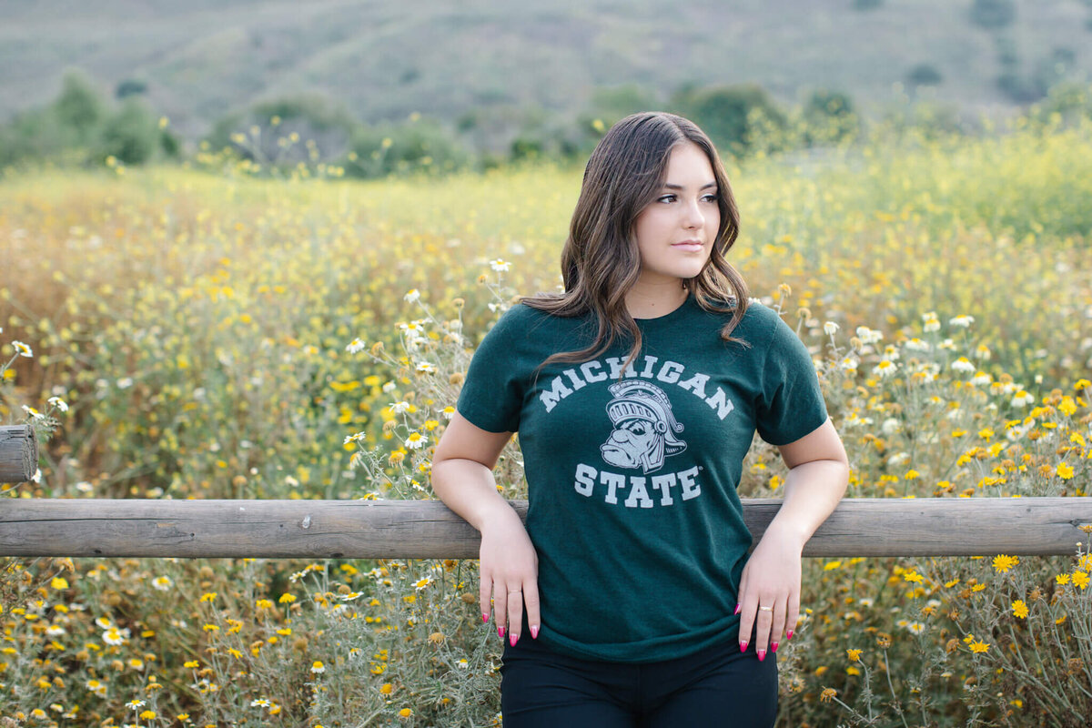 A young woman with brown hair, wearing a green T-shirt that says Michigan State, leaning her elbows against a fence and looking to her left in front of a field of yellow flowers.