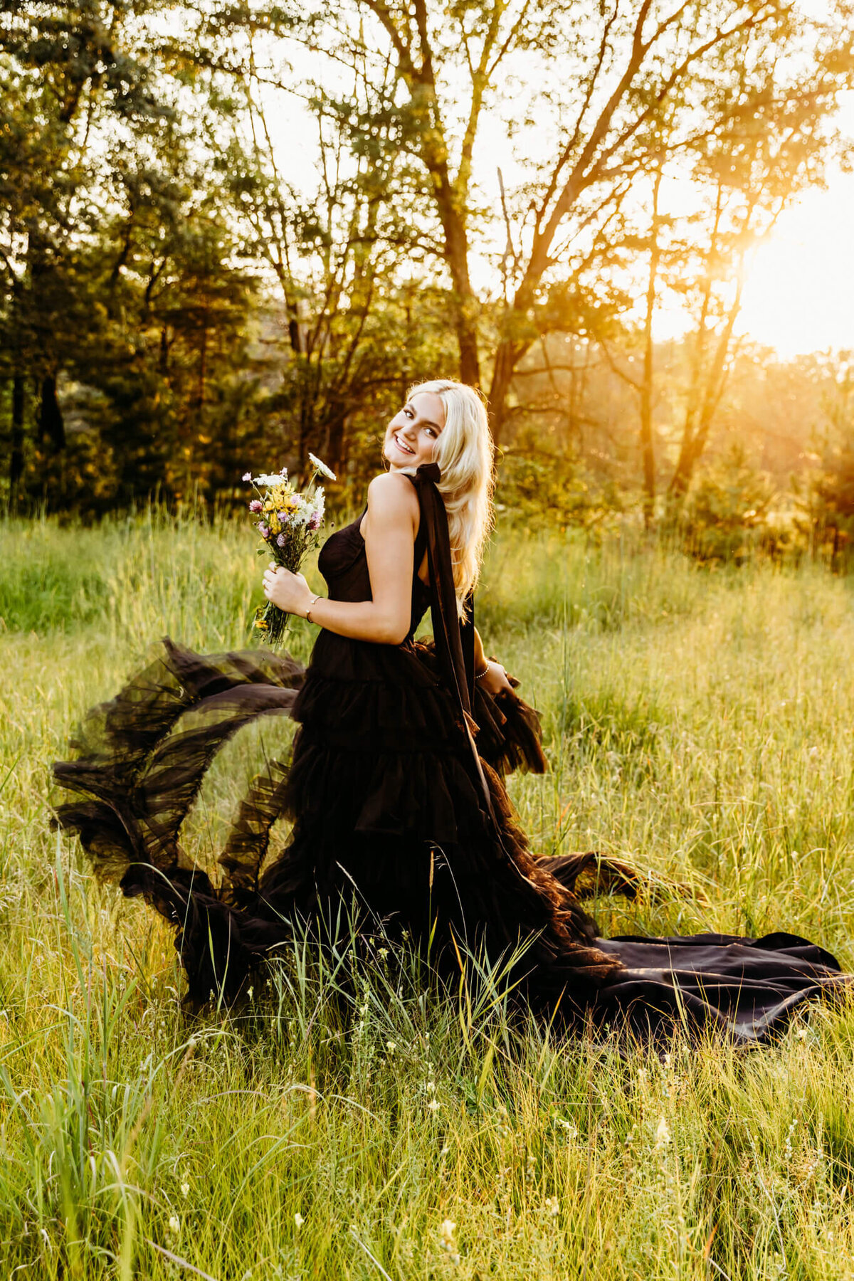 teenage girl in a large black gown spinning in the middle of a field as she holds flowers and looks over her shoulder.