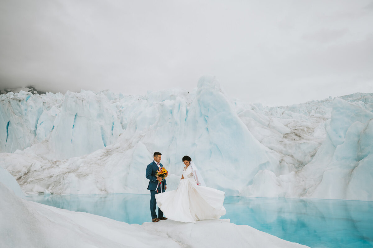 couple on their elopement day standing on a glacier in front of a blue pool while the bride twirls her dress