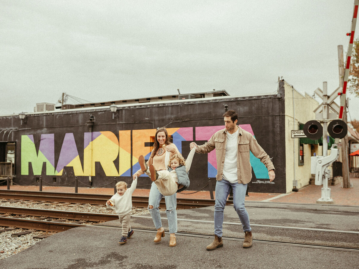 A family walks across the street swinging their toddler over the train tracks by the Marietta mural. 