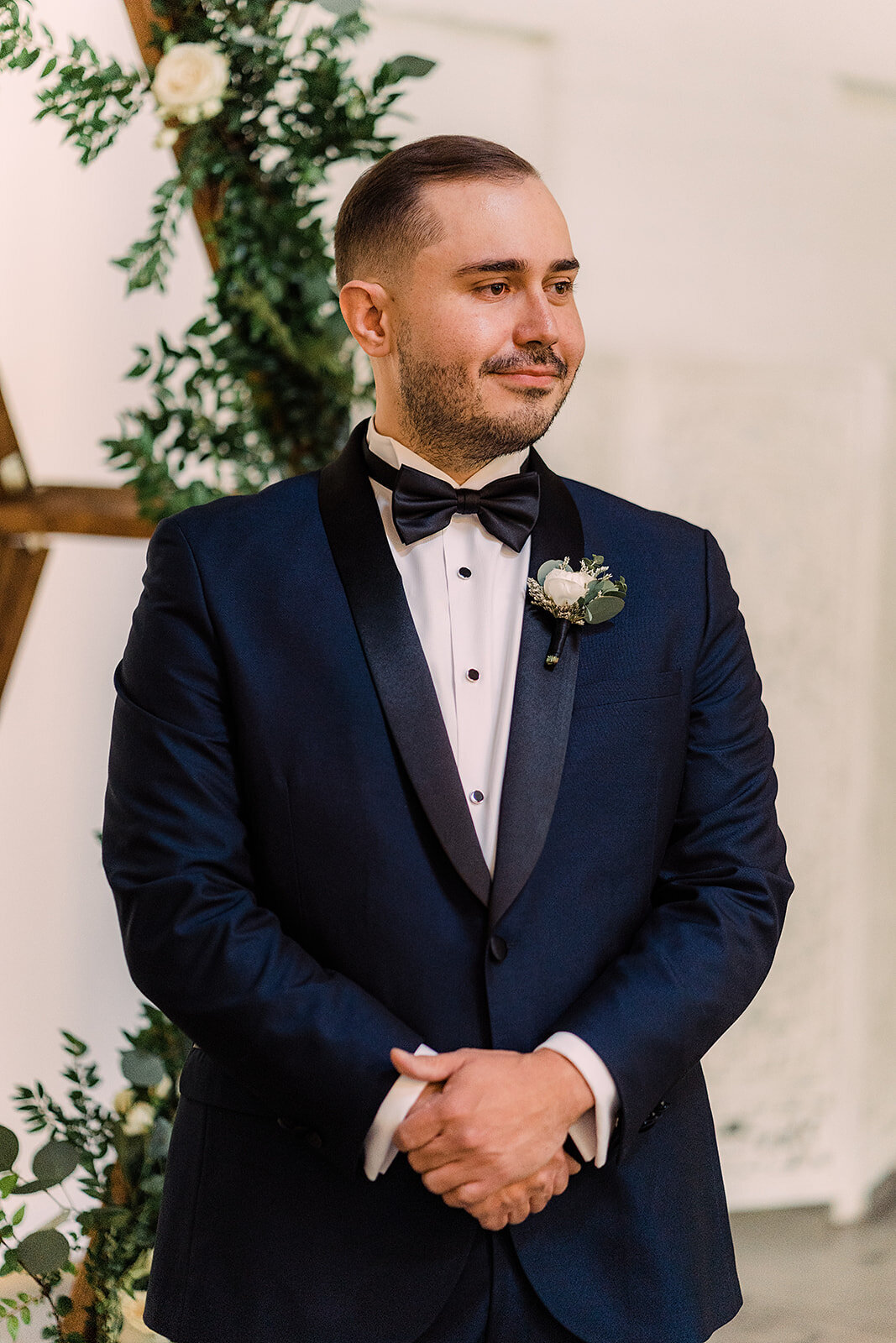 Groom stands at the altar in a sharp navy tuxedo, hands clasped and expression serene, awaiting his bride with anticipation.