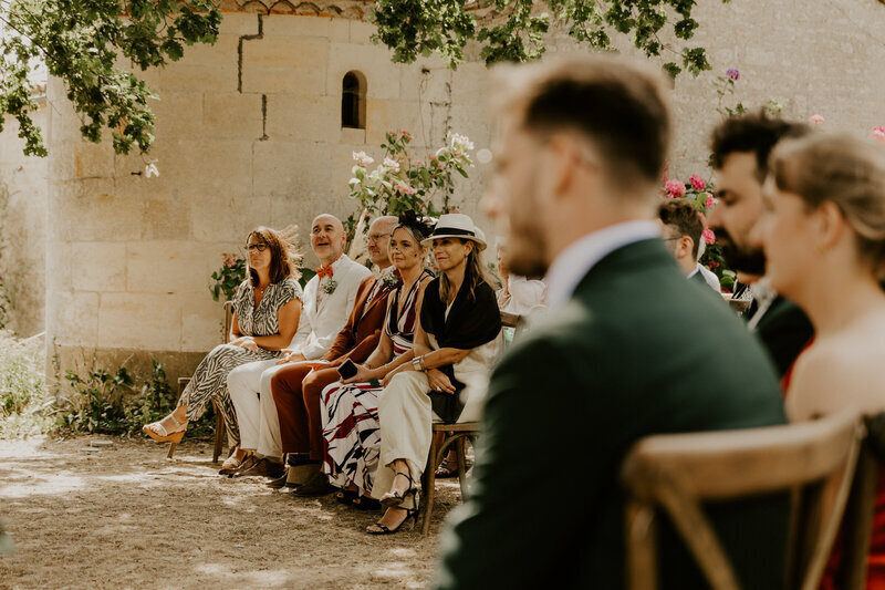 Invités assis sur des chaises en bois devant un bâtiment en pierres pendant la cérémonie laïque. Photo prise par Laura, photographe mariage en vendée.