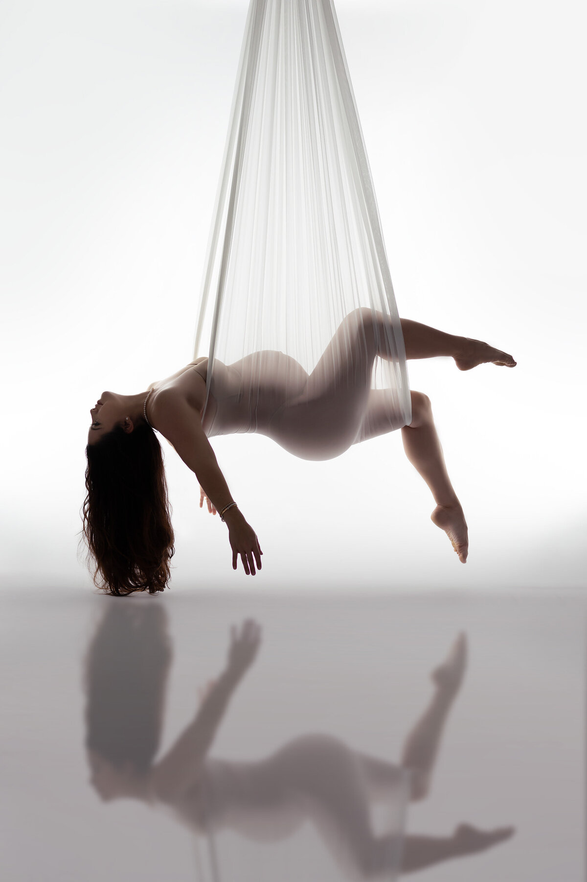 A unique silhouette of a pregnant woman suspended from sheer white aerial silks. You can see her beautiful curves and belly and her legs are bent with toes pointed.