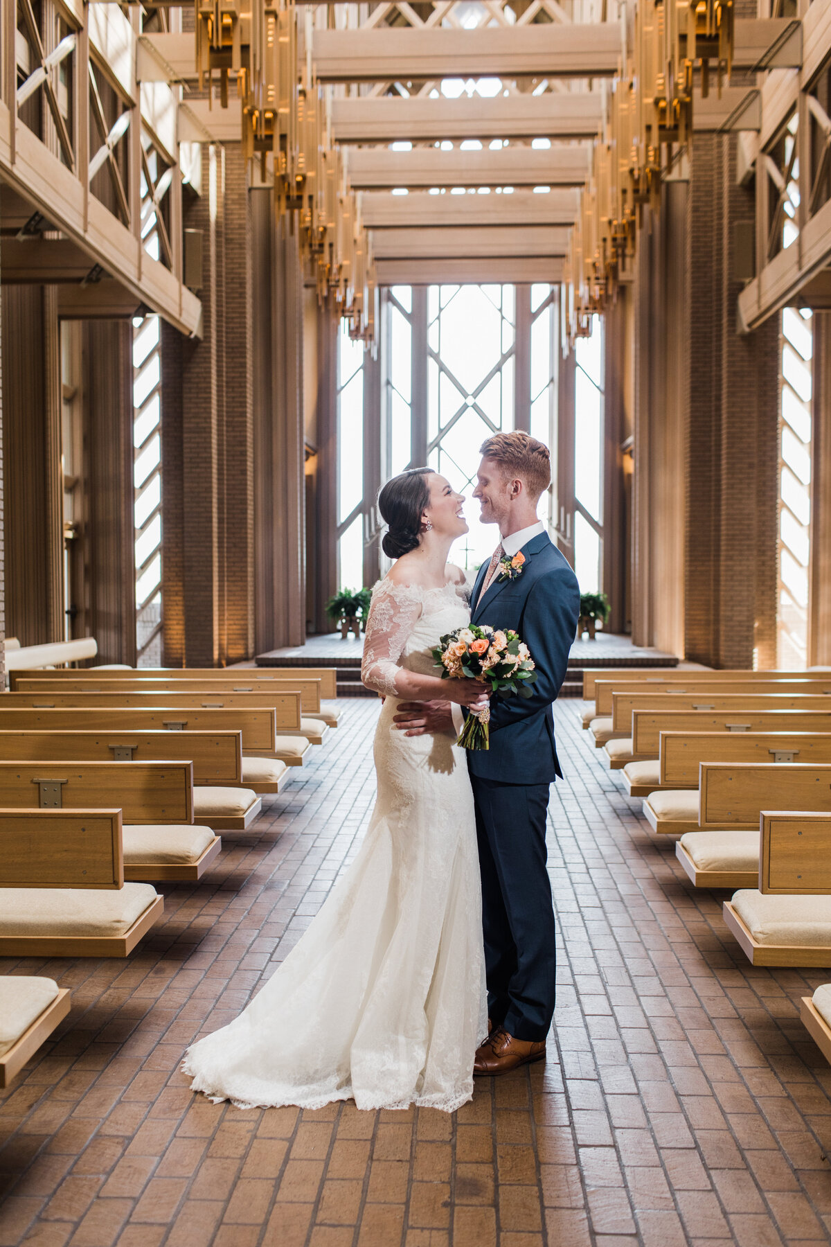 A portrait of a bride and groom holding each other close and smiling in the chapel portion of the Marty Leonard Community Chapel in Fort Worth, Texas. The bride is on the left and is wearing a long sleeve, intricate, white dress and is holding a bouquet. The groom is on the right and is wearing a blue suit with a boutonniere.