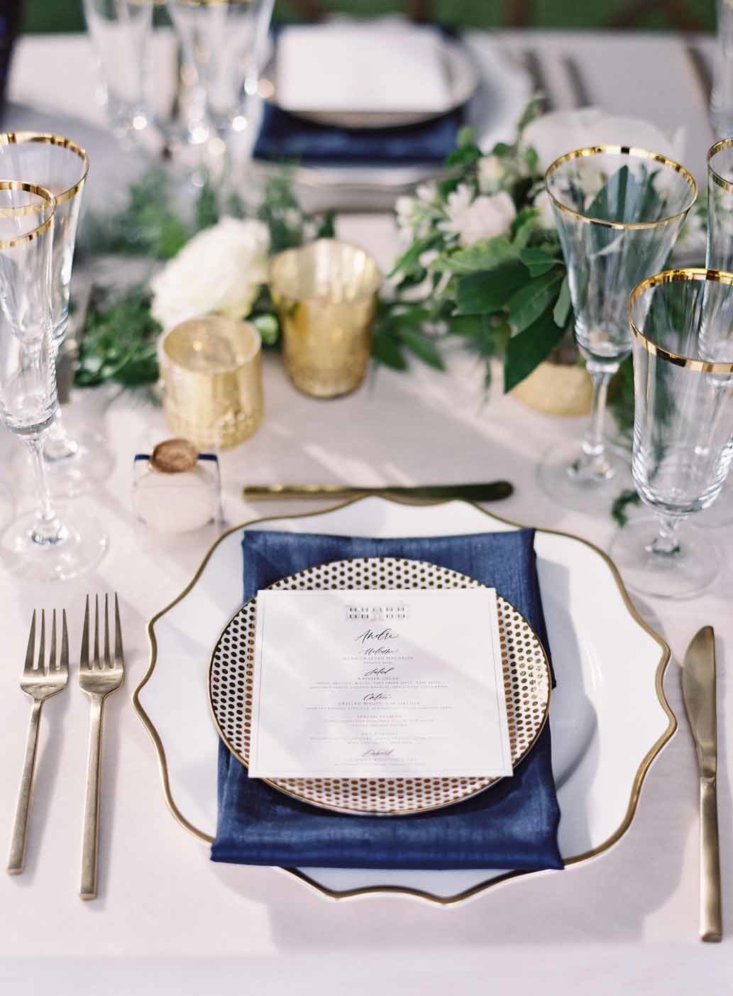 place setting for wedding with gold rimmed plates, gold flatware, navy napkin, gold votives, and greenery centerpiece