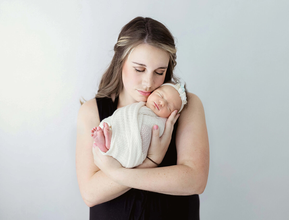 Mom with soft expression holding baby girl swaddled in white - classic newborn studio portrait  Maple Grove
