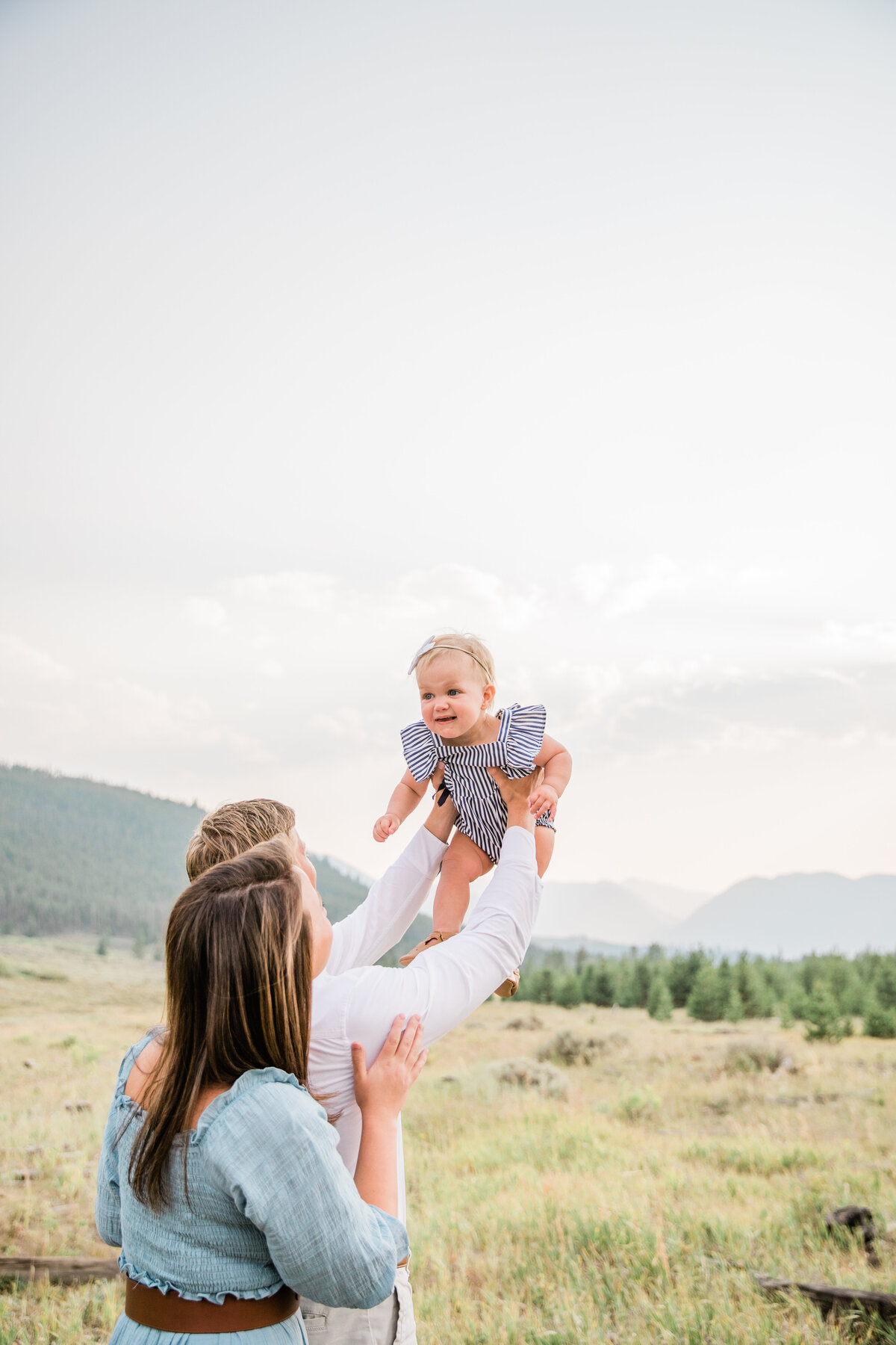 Two parents are standing close together while they hold their young baby high above their heads in a playful swing. They are in a grassy meadow with mountains behind them.