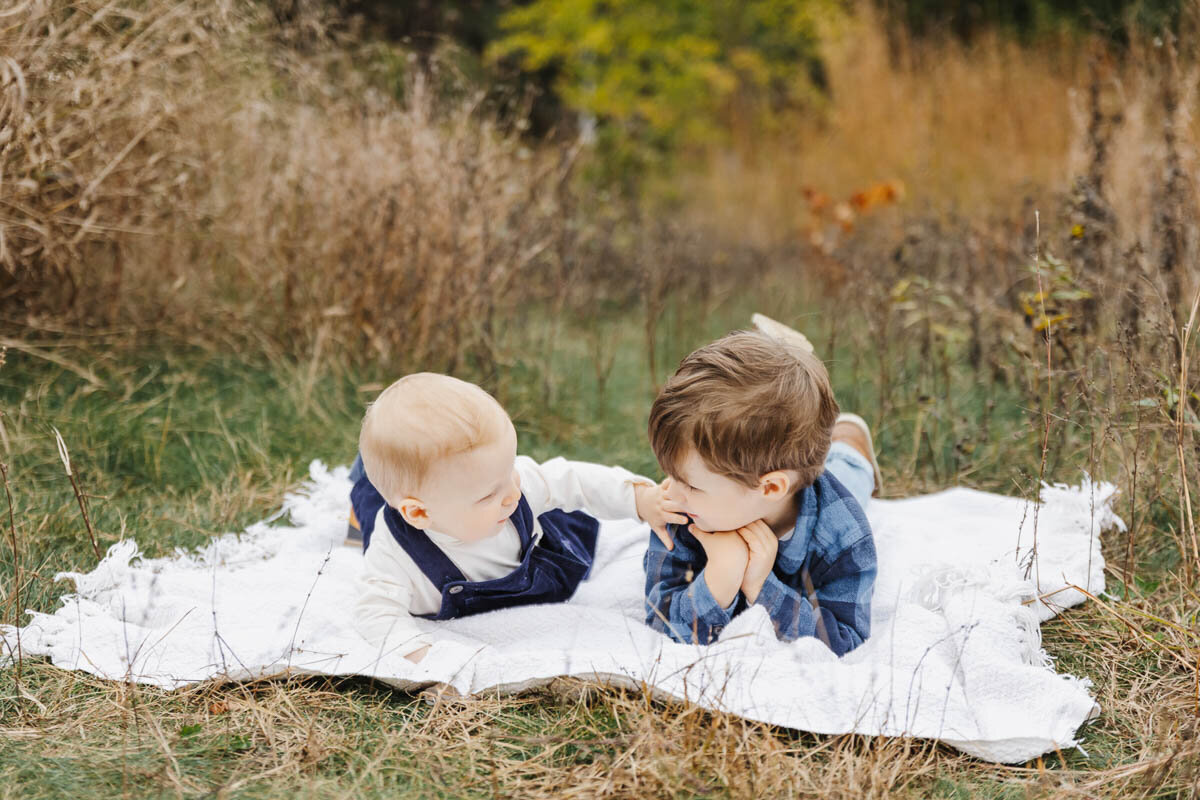 Two young brothers enjoying a giggle on a white blanket nestled in tall grass. Captured by Toronto Family Photographer Claire Binks