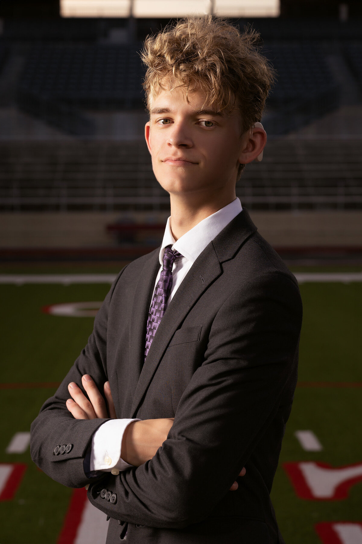 A senior in a suit stands confidently with his arms crossed on the football field at Grapevine High School. The stadium is in the background as part of this dramatic Dallas senior photography session.