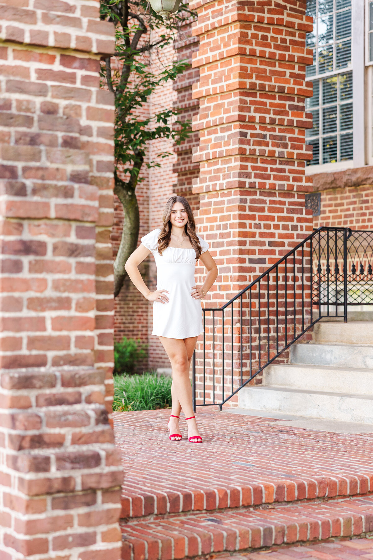 A person in a white dress and red shoes stands confidently on a brick patio framed by brick pillars, evoking the artistry of a senior portrait photographer in Fayetteville, NC. With hands on hips and a bright smile, they grace the scene where steps lead to a building with black railings and windows.