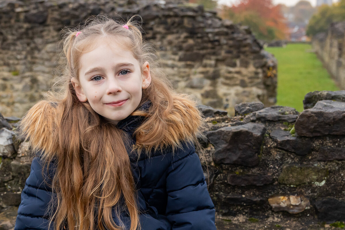 Young girl with long hair and a blue jacket smiling, sitting in front of an old stone wall with a grassy field in the background.